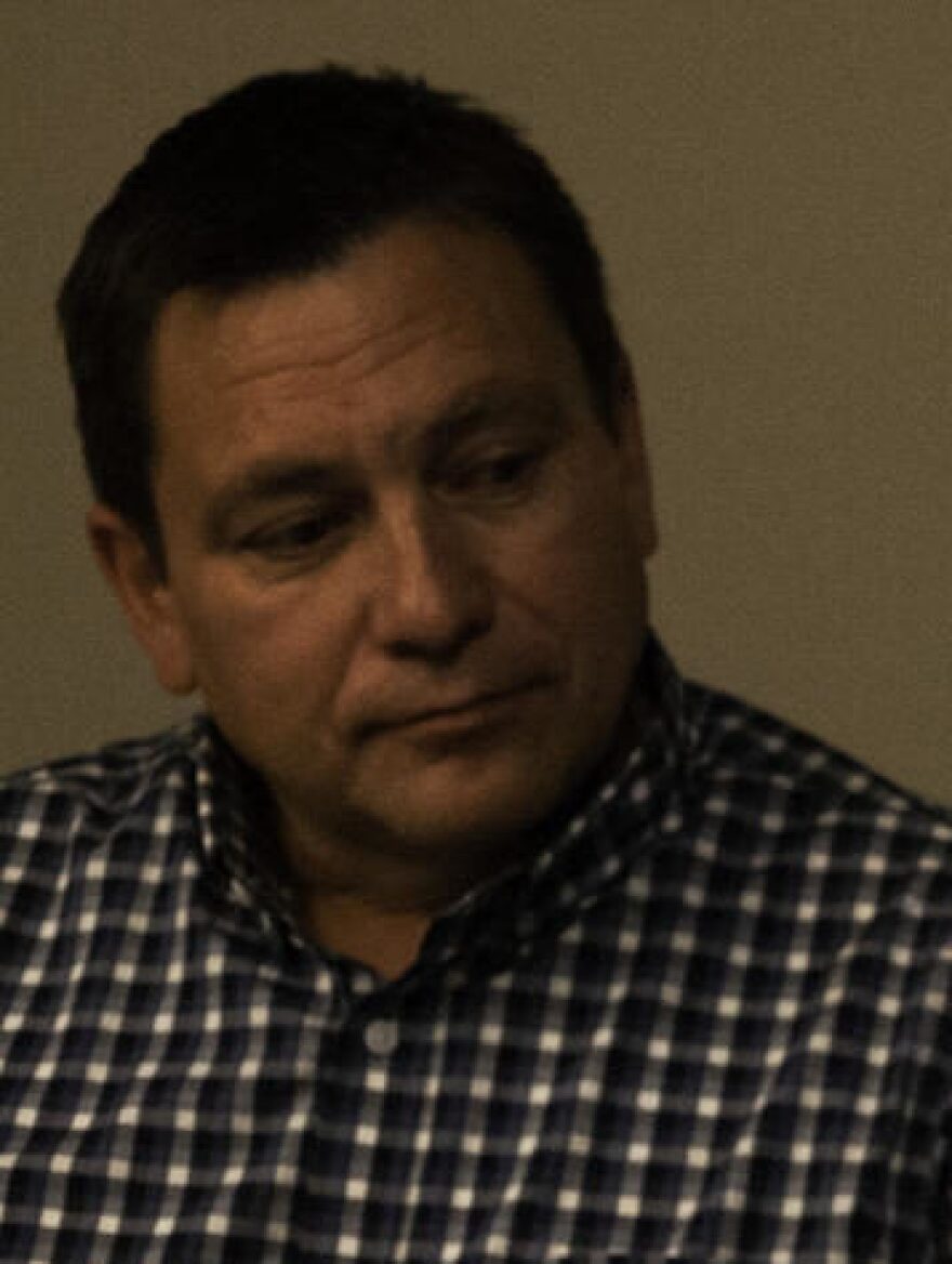 A white man with greying curly hair and a pinstripe button up shirt speaks with his arms crossed at a table. Another man listens in teh background