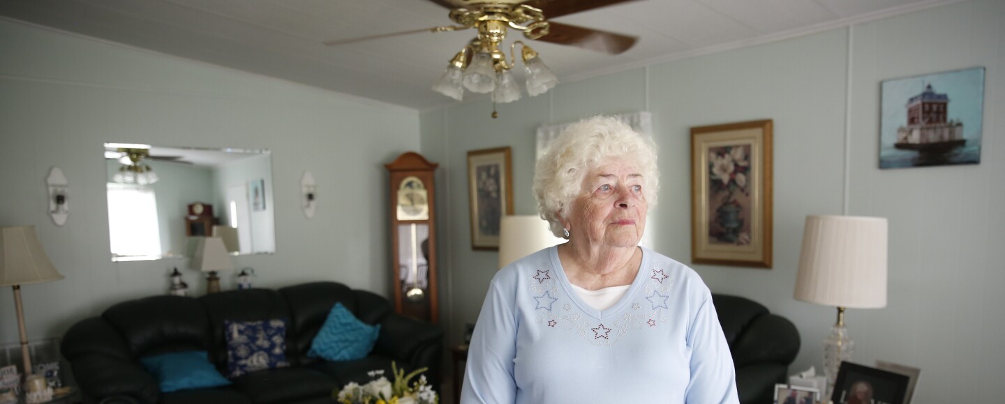 An elderly woman stands in front of her living room with a couch and other decorations in the background. 