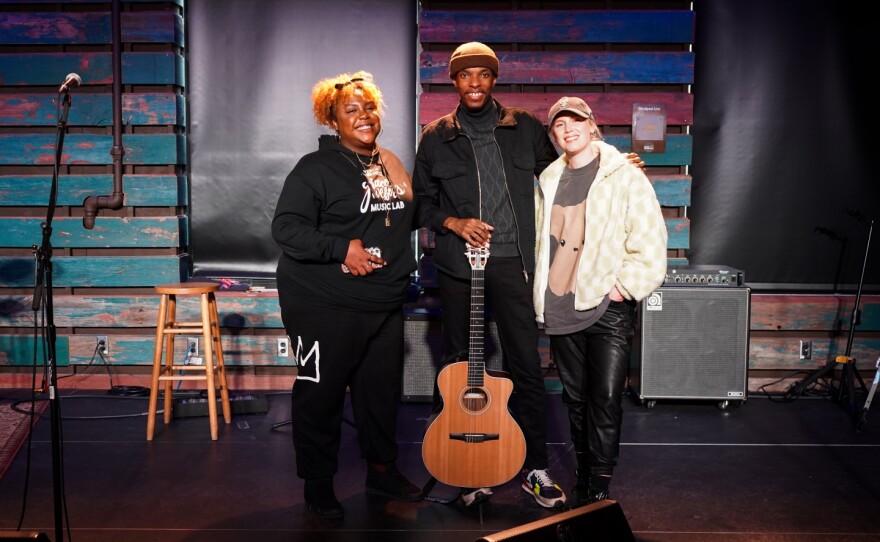 A man holding an acoustic guitar stands between two women as all three smile at the camera.