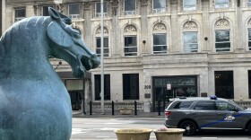 A picture taken of the Lexington Fayette Government Center from across the street. In the foreground on the left-hand side is the bust of a metal horse statue, its mouth open. A Kentucky State Police SUV passes on the road. The building at 200 West Vine Street is an early 20th century building constructed of warm, white stone blocks. 