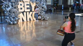 Inside a large ornate building (Union Station in Kansas City) a woman kneels to take a photograph of another woman posing with a large, lighted sign that reads "BEY INKC." The sign is surrounded by silver balloons of various shapes and styles.