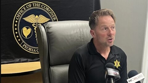 Peoria County Coroner Jamie Harwood speaks into media microphones with a banner behind his back at a 2024 news conference in his office.