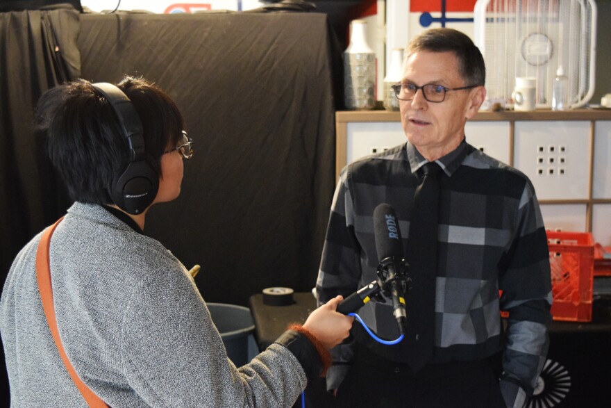 a woman with short black hair and glasses holds a mic and interviews a man in glasses and a tie.