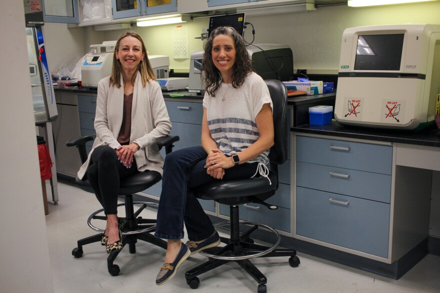 Two women sitting on rolling chairs pose for a photo in a scientific laboratory. 
