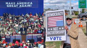 Left: President Trump at a podium on a rally stage. Right: a demonstrator with a sign that says "Democracy not dictatorship - Vote." In the background is a gas sign showing $3.99 a gallon. 