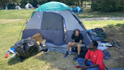 Two men sit outdoors near a camping tent. Behind them are other tents near a wooded area.