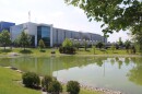 A photo of a data center from the street nearby. Behind a fence and across a green pond, fans on the roof of the center exhaust a dark smoke.