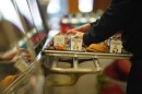Sweet potato fries, milk cartons and a chicken sandwich sit on a lunch tray on top metal rungs in the foreground. In the background, a pair of hands touches a meal on the tray in the background.