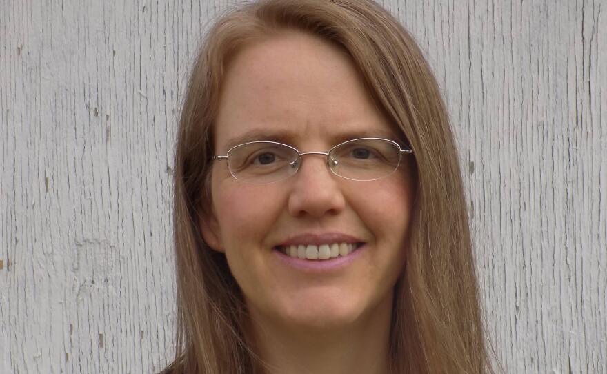 A smiling woman with dark blonde hair, wearing glasses, a pearl necklace, and a blue and pink floral top