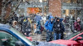 Snow falls as cars and people move across a street.