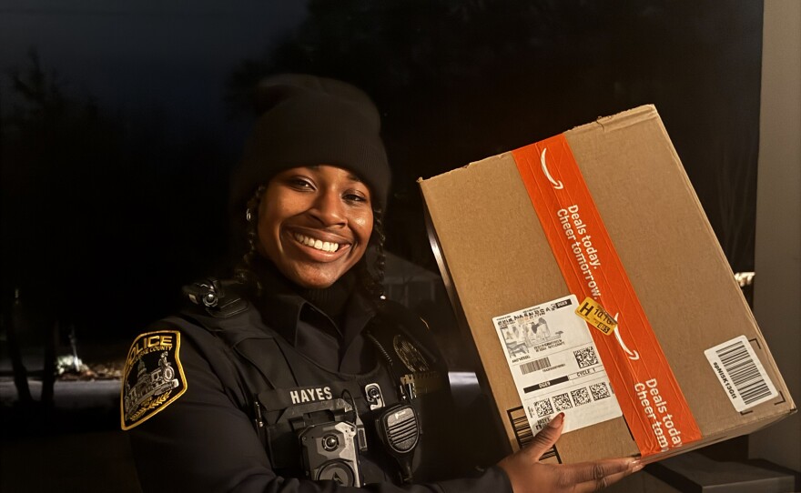 A police officer smiles while holding a large Amazon box on a residential porch at night. 