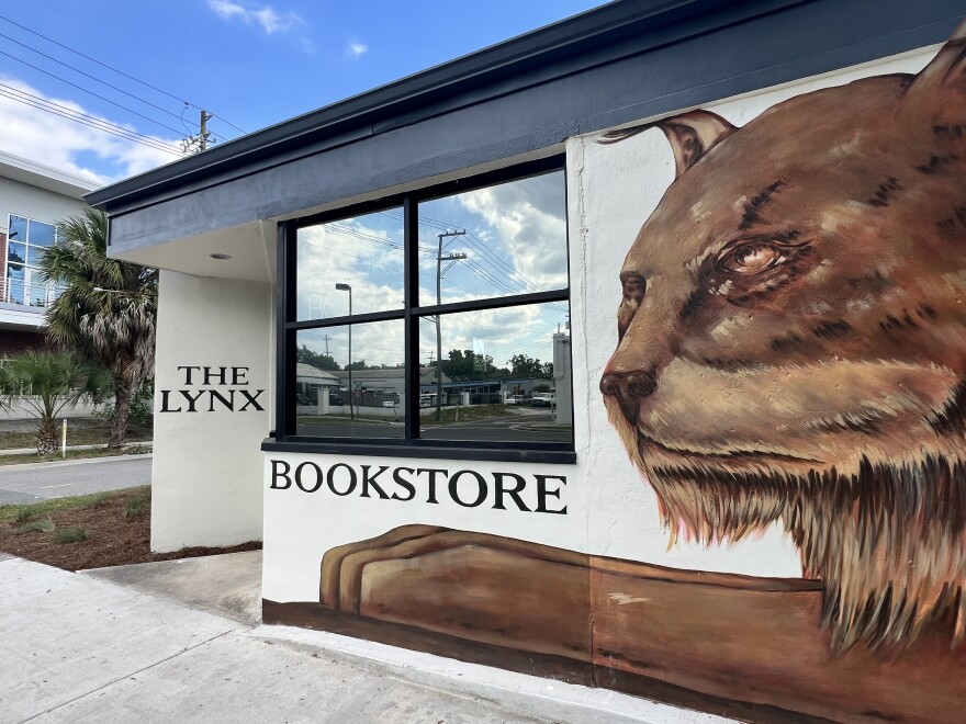 A white, one-story building with a window. Black capitalized serif letters on two of the building's walls read "The Lynx Bookstore." Next to the window is a large painting of a brown lynx's face and paw.