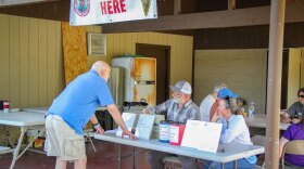 Two people sit at a folding table. A man in a blue shirt and light brown shorts stands in front of the table. A "register here" banner hangs above the table.
