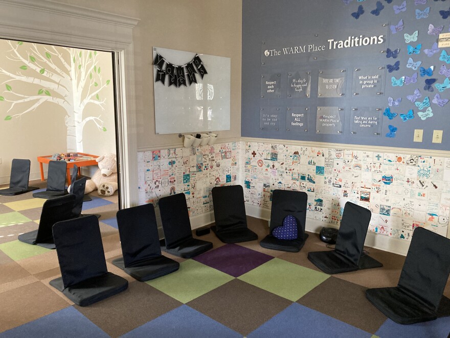 A rug with colorful squares is surrounded by black chairs, with walls covered by children's artwork.