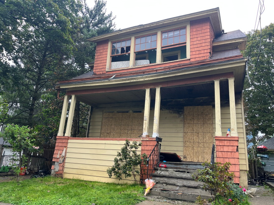 This is a photo of the red and orange house where the fire broke out. Two of the the three front-facing windows on the second floor are broken. The third, on the right, was left open. There's some charred pieces in the driveway. The door and front floor windows are boarded up. You can see fire damage on the exterior wall above the door. 