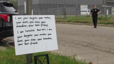A sign sits on a street that says "I am your daughter, your mother, your aunt, your sister, your grandma, your best friend. I am homeless. But still I am our daughter,  your mother, your aunt, your sister, your grandma, and your best friend." A police officer stands in the background. 