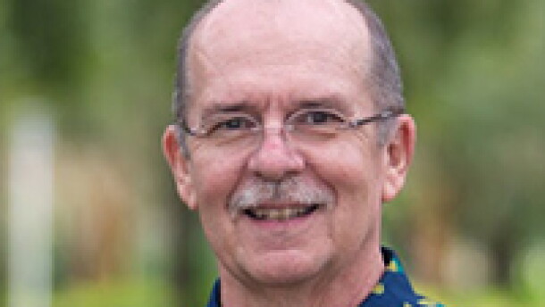 Bald man with a gray mustache and glasses wearing a blue short-sleeve button-down shirt and smiling into the camera