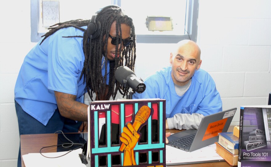 Two student DJs pose for the camera behind a desk. On the left, a young black man with long braids and dark sunglasses leans over a microphone. On the right, a young Latino man with a shaved head smiles at the camera.