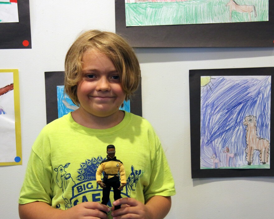 J.R. Patterson stands in front of a wall of art. He has medium length dirty blond hair, is wearing a neon yellow shirt and is holding a Star Trek action figure.