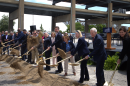 A group of people in dress clothes shovel dirt next to a large steel bridge