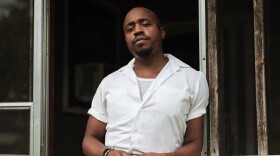 A man in a white button-down shirt with short sleeves stands in the entrywayof a dilapidated porch.