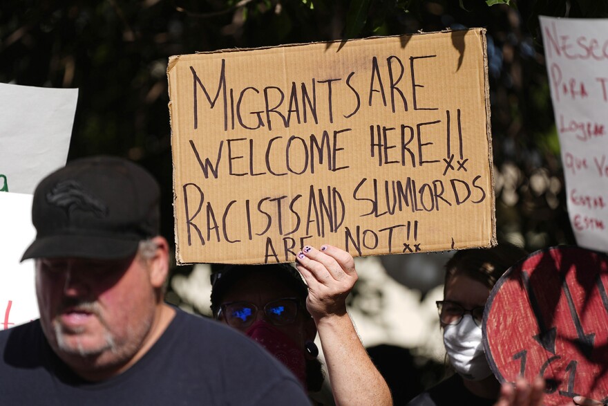 A person holds up a sign written on cardboard that says "Migrants are welcome here!! Racists and slumlords are not!!"