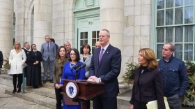 Massachusetts Governor Charlie Baker stands at a podium in front of a marble building with a row of people standing on either side of him