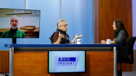 A man and woman sit at a desk speaking to each other, while a third person watches from a nearby television monitor.