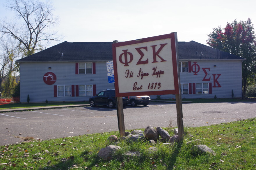 A large grey building sits in the background, with a sign in the foreground marking it as the Phi Sigma Kappa fraternity, with matching Greek letters. These letters can also be seen on the side of the building. 