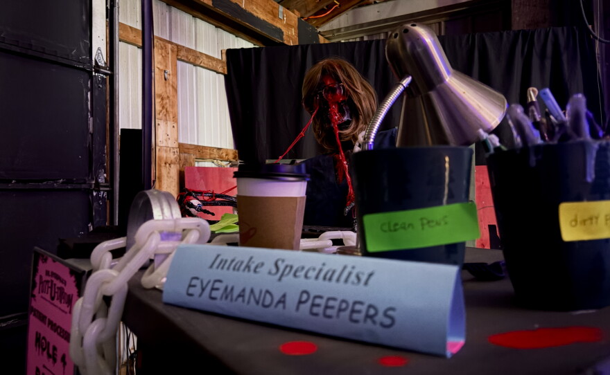 A desk full of pens, paper and chains. Behind the desk is a statue called Eyemanda.