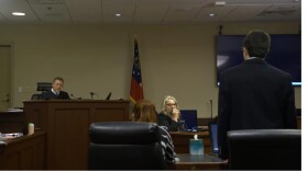 A courtroom scene shows a judge seated at the bench while an attorney stands facing him and other participants sit nearby.