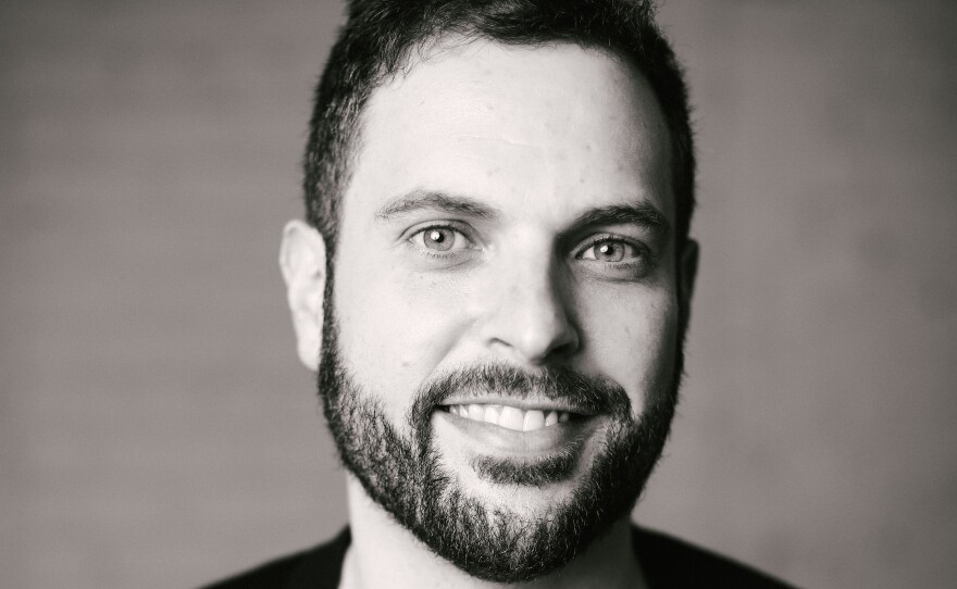 A man with dark hair and a dark beard wears a cardigan and a t-shirt.