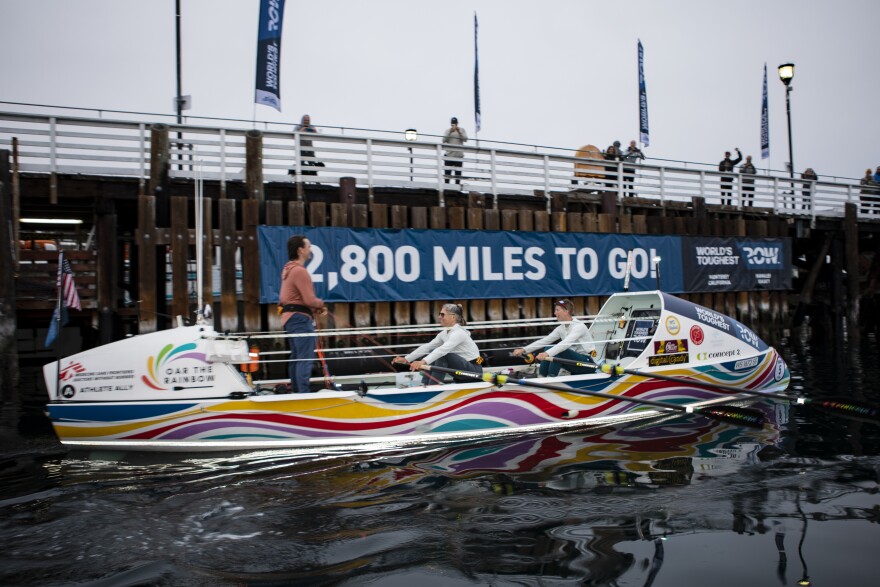 Two people wearing white longsleeve shirts row a boat in the ocean and face one person who is standing up at the stern of the boat wearing an orange longsleeve shirt. The boat is painted in rainbow-colored waves and there is a wooden fishing pier in the background. 