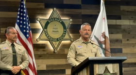 A man in a sheriff's uniform stands behind a podium next to another man in the same uniform. 