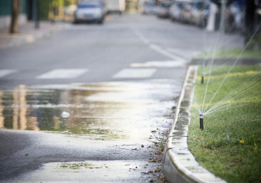 A photo of a garden sprinkler in action near a grass covered traffic island, with water on the sidewalk.