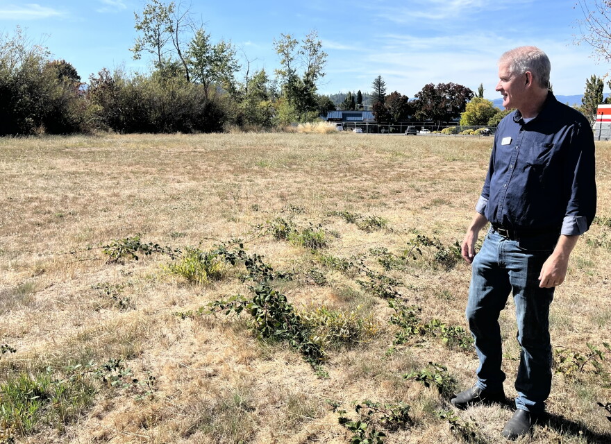 A middle aged man wearing jeans and a dark blue button down is standing in a dry grass field, gazing off to the left. 