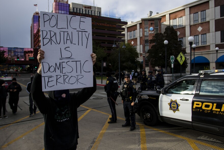 A person dressed in all black holds a piece of white cardboard that reads, “Police brutality is domestic terror.” Behind the person is a Reno Police Department SUV and police officers.