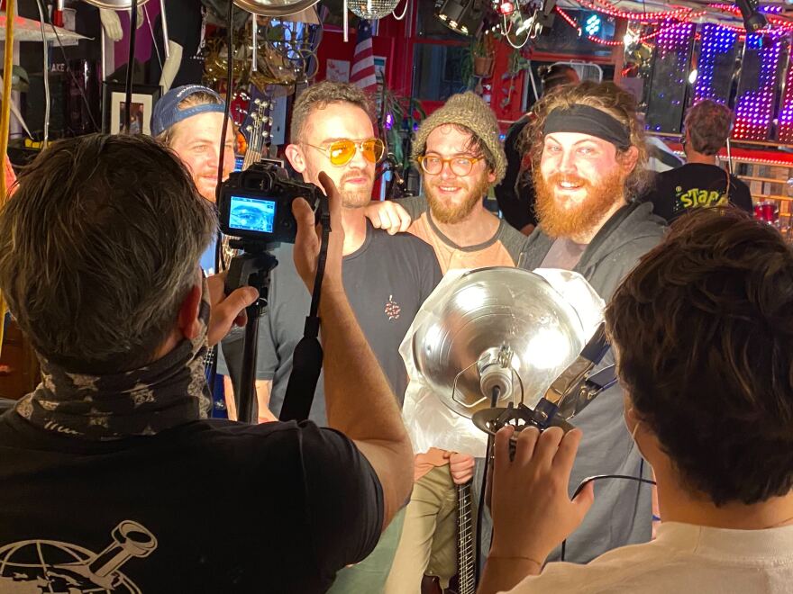 Four men are lined up in front of two other men, one holding a light and the other a camera