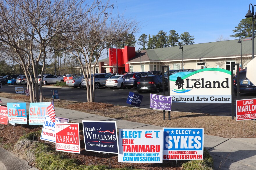 A group of colorful political signs staked in the ground in front of the polling location at the Leland Cultural Arts Center, which is a tan one-story building with a tall red pillar. It's a sunny afternoon.