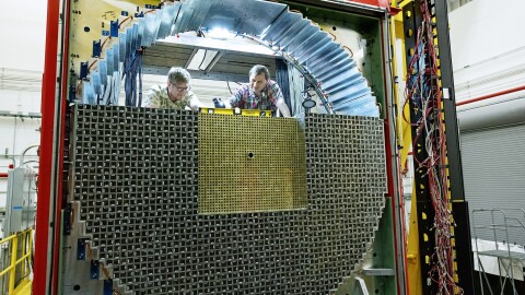 Two men are perched atop a large circular object looking down at something in the machine. Wires are wrapped around the right side of the machine, and small squares fill half the circle of the machine, almost like a beehive. 