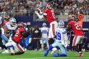 football action with player #0 in red jersey and pewter helmet and white pants jumping to catch the ball. teammates are on each side along with opposing players in white jerseys and gray pants and helmet