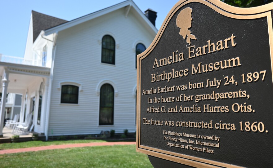 A bronze sign that reads "Amelia Earhart Birthplace Museum" and some information about Earhart stands in front of a white, wooden house.