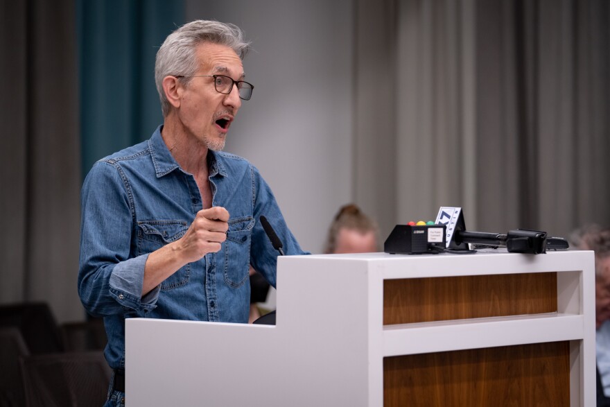 Ken Zarifis, the president of Education Austin, is wearing a blue denim shirt and he is standing behind a white and wood-colored podium while talking to Austin ISD school board members who are not pictured.