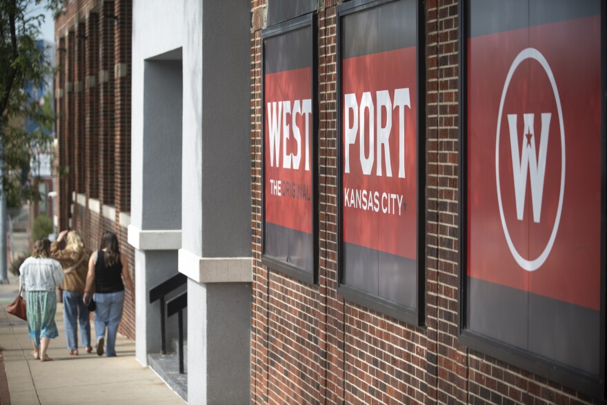 Exterior photo shows three people walking on a sidewalk away from the camera. A large brick building at right has a large painted sign that reads "Westport, the Original Kansas City."