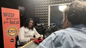 African American woman sits with headphones on near microphone in front of WUSF Your Spot for Local sign. A man's back with headphones over his ears is also seen sitting across from her next to another microphone.