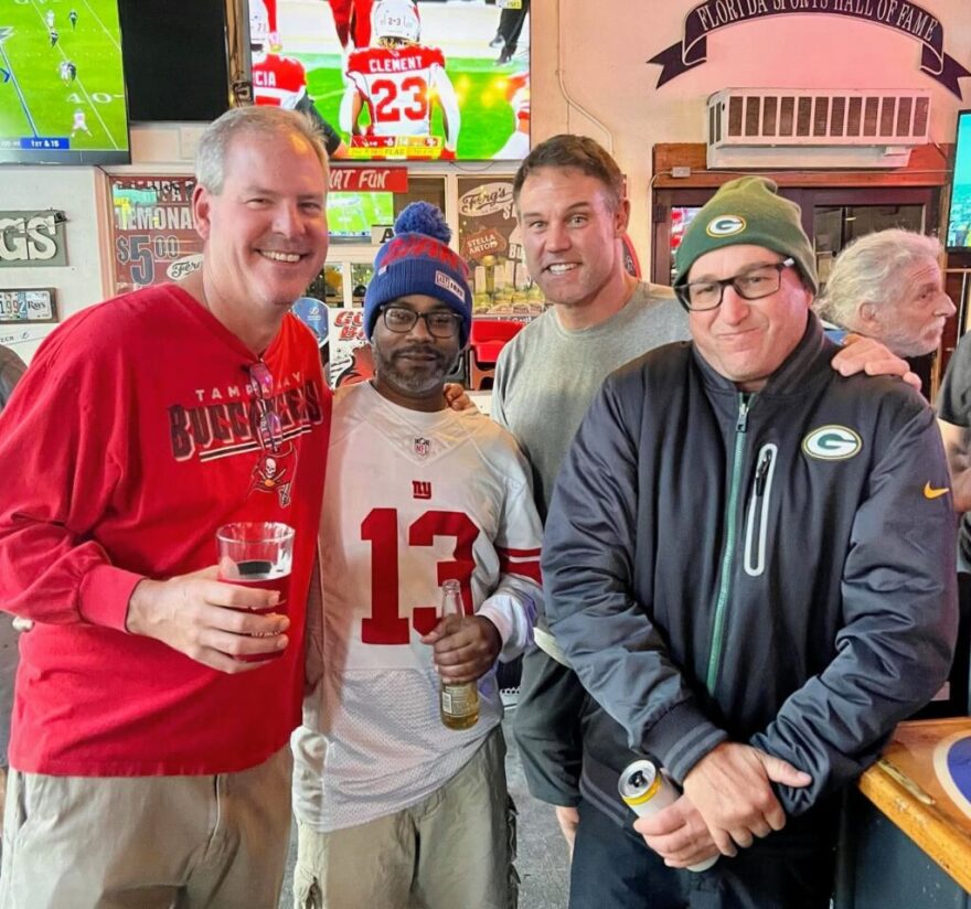 Four men wearing football shirts and standing next to each other in a bar, smiling into the camera