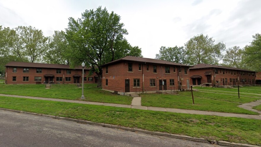 Three two-story red brick apartment buildings with brown roofs sit on a grassy lawn, surrounded by trees and sidewalks, under a cloudy sky.