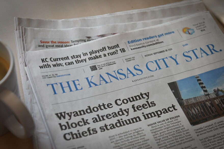 A newspaper sits flat on a table with a coffee cup nearby. The paper reads "The Kansas City Star" across the nameplate.