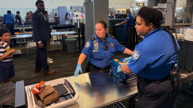 Two TSA officers dressed in crisp blue uniforms scan luggage and carry-on items at a security checkpoint in Tampa International Airport. Travelers can be seen going through security in the background.