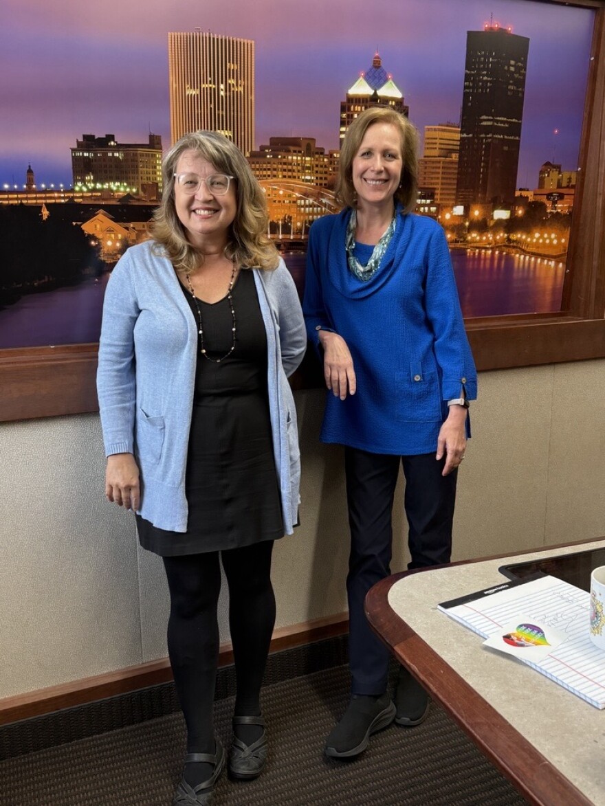 Two smiling women stand in front of a city backdrop in a radio talk studio: a woman at left has long blonde hair and is wearing glasses, a blue cardigan, black dress, black tights and black shoes; a woman at right has long blonde hair and is wearing a bright blue sweater, blue pants and black shoes.